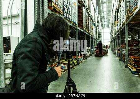 Un videografo si vede dietro una fotocamera reflex professionale montata su un treppiede all'interno di un magazzino, filmare i supporti promozionali per l'azienda, copiare lo spazio a destra. Foto Stock