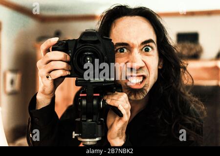 Un ritratto di fronte da vicino di un fotografo caucasico con un'espressione del viso divertente e scricchiolante, dietro una fotocamera reflex digitale professionale e un treppiede. Foto Stock