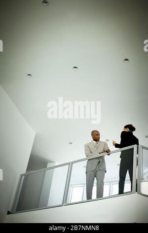 Un uomo d'affari medio-adulto che si appoggia su un banister all'interno di un edificio d'ufficio che parla con una collega femminile. Foto Stock