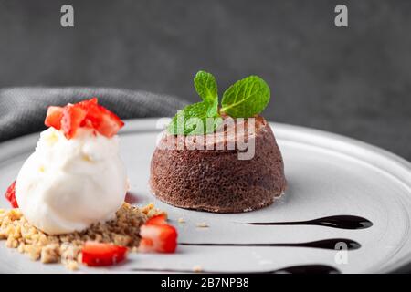 Piatto bianco con torta al cioccolato fondente con gelato e fragola su sfondo scuro decorato menta e sciroppo, vista dall'alto, orizzontale Foto Stock