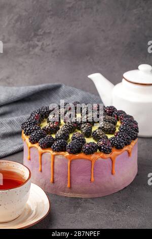 Torta intera ricoperta di crema viola, more fresche, caramello, scorre sui lati, teiera e tazza da tè su superficie grigia, verticale Foto Stock