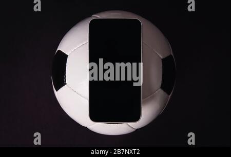 Concetto di scommesse sul calcio dal vivo, trasmissione online, display smartphone vuoto sulla classica palla di calcio isolato su sfondo nero. Sport gioco d'azzardo copia sp Foto Stock