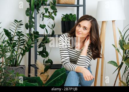 Bella ragazza siede tra le piante domestiche, lei in una giacca a righe e jeans Foto Stock