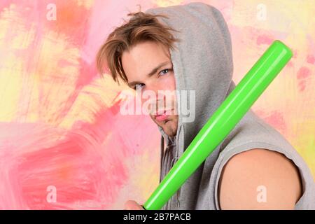 Il ragazzo in felpa con cappuccio grigio senza maniche tiene il pipistrello verde brillante. Gangster di strada con volto concentrato pronto a colpire. Uomo con cappuccio sulla testa su sfondo colorato. Concetto di vita di strada, pericolo e crimine Foto Stock