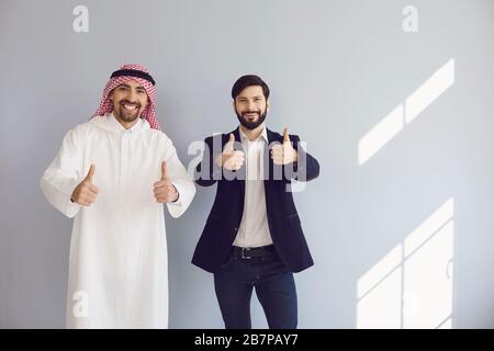 Uomini d'affari arabi ed europei mostrano il pollice in piedi sorridente sullo sfondo grigio. Foto Stock