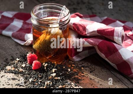 Sacchetto di tè fatto in casa che si macina in un vaso di mason, con tè e lamponi versati davanti, e un panno rosso-e-bianco controllato dietro. Foto Stock