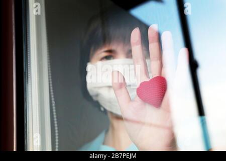 Quarantena durante l'epidemia di coronavirus COVID-19, paziente infetto. Donna malata in una maschera medica del viso guarda attraverso il vetro della finestra con il cuore rosso Foto Stock