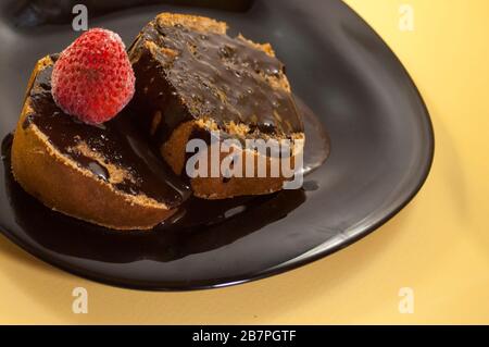 raffigurazione di due pezzi di torta ricoperta di cioccolato con diverse fragole sulla parte superiore in piatto nero su sfondo giallo tavolo Foto Stock
