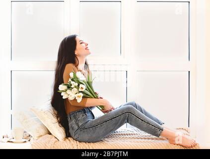 Vista laterale di una giovane donna soddisfatta in un vestito informale che tiene bouquet di tulipani bianchi e sorridente mentre si siede sul davanzale in una camera accogliente a casa Foto Stock