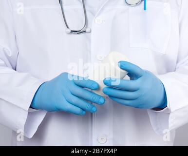 medico maschile in un camice medico bianco e guanti in lattice blu contiene un vaso di plastica bianca di pillole, primo piano Foto Stock