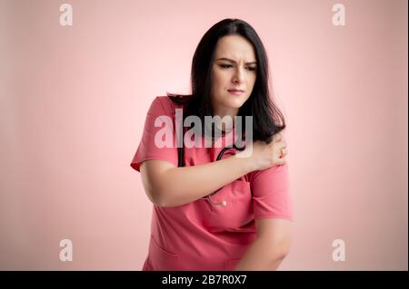 Ritratto di bella donna medico con stetoscopio che indossa scrub rosa, ha dolore alle spalle in posa su un rosa isolato backround. Foto Stock