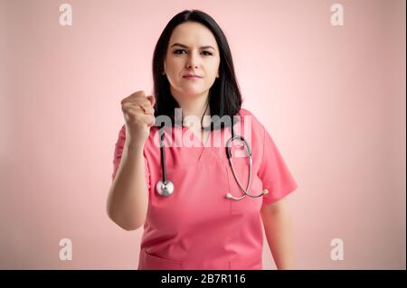 Ritratto di bella donna dottore con stetoscopio con scrub rosa, mostrando pugno posando su un dorso isolato rosa. Foto Stock