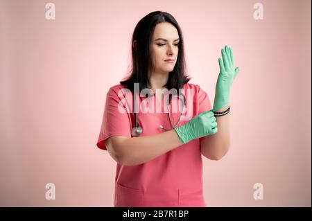 Ritratto di bella donna medico con stetoscopio che indossa scrub rosa, prende guanti medici sulle sue mani in posa su un dorso isolato rosa. Foto Stock