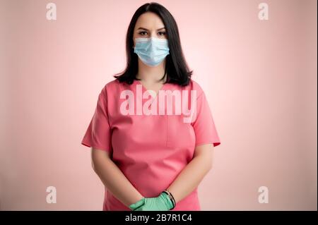 Ritratto di bella donna medico con stetoscopio con scrub rosa, con maschera protettiva in posa su un dorso isolato rosa. Foto Stock