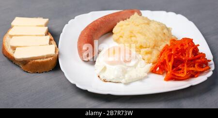 pranzo gustoso e gustoso. salsiccia all'uovo e patate tostate con un panino cheesy su un piatto bianco. primo piano su sfondo grigio. Foto Stock