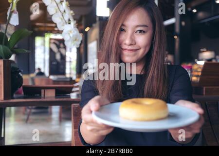 Una bella donna asiatica che tiene e mostra un piatto di ciambella con sentirsi felice e buon stile di vita nel moderno caffè Foto Stock