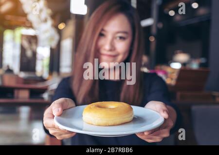 Una bella donna asiatica che tiene e mostra un piatto di ciambella con sentirsi felice e buon stile di vita nel moderno caffè Foto Stock