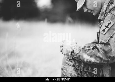 Colpo in scala di grigi di un giovane soldato che prega mentre si inginocchiava un'erba asciutta Foto Stock