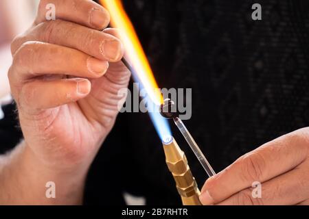 Le mani dell'uomo artigianale che fa un soggetto di vetro di mestieri. Produttore di vetro che lavora vetro fuso con una torcia a fiamma. Primo piano della fiamma dalla torcia a soffiante Foto Stock