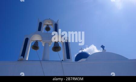 Torre della Chiesa Blu e Bianca a Santorini Grecia retroilluminata con tre campane a cupola e due croci con nuvole bianche in un chiaro cielo blu giorno Foto Stock