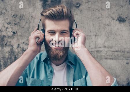 Bell'elegante ragazzo bearded rosso con un sorriso radioso e un'acconciatura alla moda perfetta sta ascoltando la musica all'aperto con grandi cuffie Foto Stock