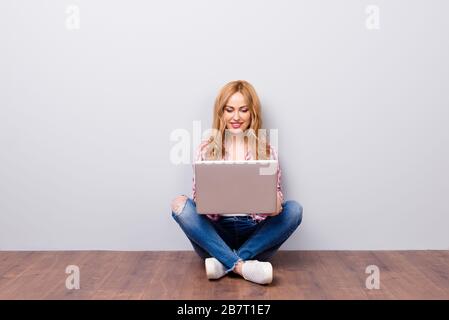 Felice giovane allegra donna in camicia a scacchi e jeans seduti sul pavimento con il laptop in ginocchio e scrivere e-mail a un amico Foto Stock