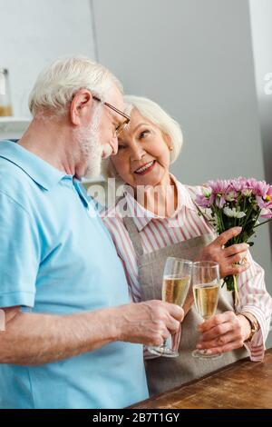 Donna anziana con bouquet sorridente al marito mentre si aggattava con champagne in cucina Foto Stock