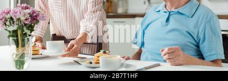 Vista tagliata della donna mettere caffè e pancake sul tavolo da marito, shot panoramico Foto Stock