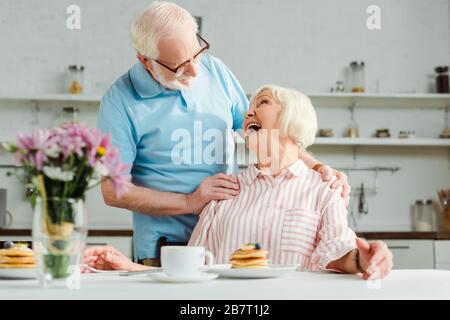Fuoco selettivo della donna anziana che ride mentre guarda il marito da caffè e pancake sul tavolo in cucina Foto Stock