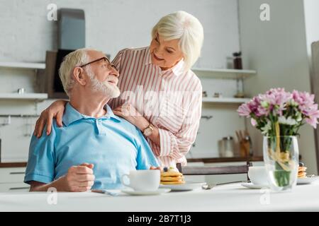 Il fuoco selettivo della donna anziana che abbracciano il marito sorridente dal caffè e dalle frittelle sulla tabella Foto Stock