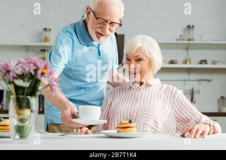 L'attenzione selettiva dell'uomo anziano che mette il caffè sul tavolo da una moglie sorridente durante la colazione Foto Stock
