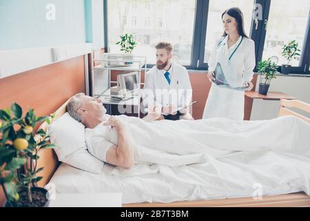 Un giovane medico e un infermiere visitano pazienti anziani presso il reparto ospedaliero moderno e luminoso. Tutti sembrano seri, discutendo i problemi della sua salute Foto Stock