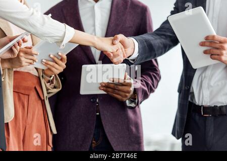 vista ritagliata di quattro colleghi multietnici che detengono tablet digitali e scuotono le mani Foto Stock