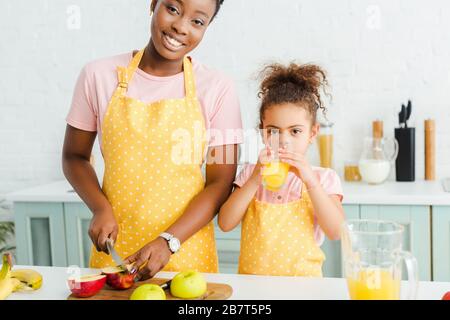 felice madre afro-americana sorridente mentre tagliando la mela vicino alla figlia bevendo succo d'arancia Foto Stock