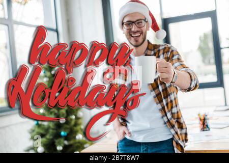 fuoco selettivo dell'uomo bearded felice in tazza di tenuta del cappello di santa vicino all'albero di natale in ufficio con illustrazione felice di festa Foto Stock