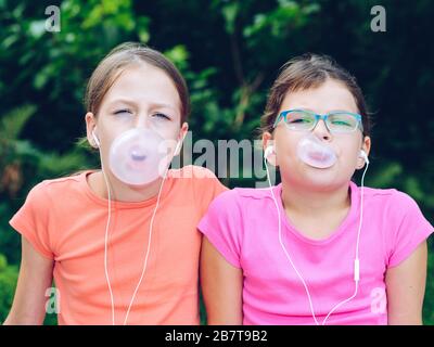 Ragazze che condividono le cuffie per ascoltare musica. Le sorelle masticano la gomma e ne fanno i palloncini. Amici felici che trascorrono il tempo insieme Foto Stock