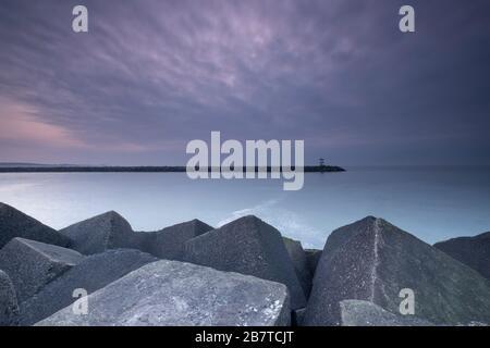 Grandi blocchi di pietra grigia con un faro in lontananza, al crepuscolo, Scheveningen, Paesi Bassi Foto Stock