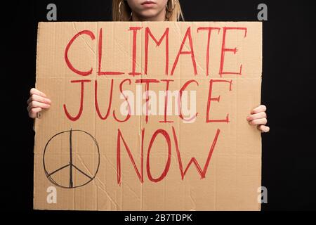 Vista parziale della donna bionda che tiene il cartello con la giustizia climatica ora scritta isolato sul nero, concetto di riscaldamento globale Foto Stock