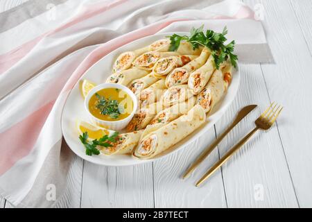 Finger food: Crepes ripieno di formaggio cremoso e salmone, prezzemolo servito su un piatto bianco su un tavolo di legno bianco con salsa di burro fuso, limone Foto Stock