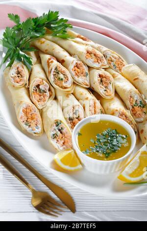 Frittelle sottili farcite con formaggio cremoso e salmone, prezzemolo servito su un piatto bianco su fondo di legno bianco con salsa di burro fuso, limone Foto Stock