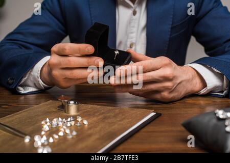 Vista ritagliata della scatola di tenuta dei monili dell'appraiser con anello dei monili vicino alle gemstones sul tavolo su sfondo grigio Foto Stock