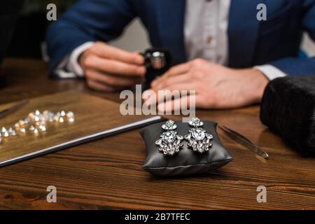 Fuoco selettivo degli orecchini sul cuscino dei monili e sull'appraiser dei monili che lavorano alla tabella in officina Foto Stock