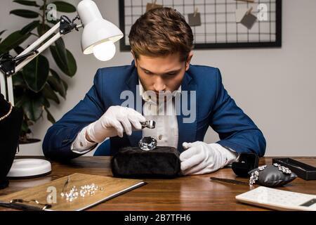 Gioielleria appraiser con lente di ingrandimento esame gemstone vicino gioielli in tavola in officina Foto Stock