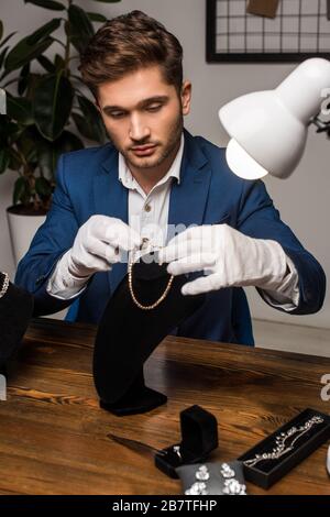 Bel gioiello appraiser in guanti che tengono collana vicino al piedistallo della collana e gioielli sul tavolo Foto Stock