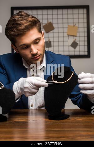 Bel gioiello appraiser tenendo pinzette vicino alla collana sul piedistallo sul tavolo in officina Foto Stock