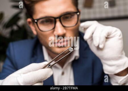 Fuoco selettivo di appraiser della gioielleria che tiene gemstone nelle pinzette mentre lavora in officina Foto Stock