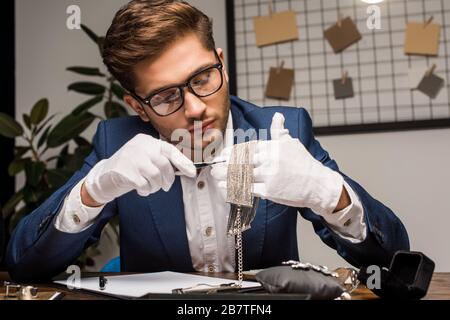 Gioielleria appraiser che tiene pinzette e collana vicino alla clipboard e gioielli sul tavolo Foto Stock