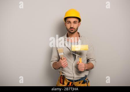 uomo lavoratore manuale in hardhat tenendo rullo di vernice e pennello su grigio Foto Stock