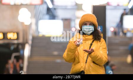 La giovane donna europea in maschera facciale monouso protettiva utilizzando antisettico nella metropolitana. Nuovo coronavirus (COVID-19). Concetto di auto sanitaria Foto Stock