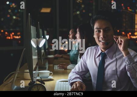 Ritratto del servizio di assistenza clienti maschile asiatico con donna d'affari sorridente e che lavora duro a tarda notte turno in ufficio, reparto call center, lavoratore A. Foto Stock